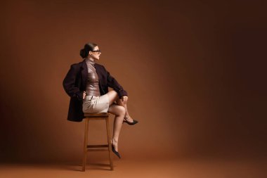 Kahverengi arka planda sandalyede şık giyinmiş genç bir kadın, mesaj için yer var.