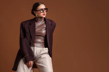 Kahverengi arka planda sandalyede şık giyinmiş genç bir kadın, mesaj için yer var.