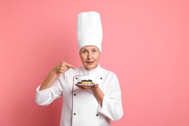 Profesyonel pasta şefi pembe arka plandaki tatlıyı işaret ediyor.