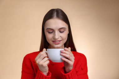 Gülümseyen kadın bej renkli arka planda fincandan kahve içiyor.