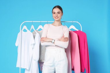 Gülümseyen moda tasarımcısı ve açık mavi arka planda giysileri olan askılar.