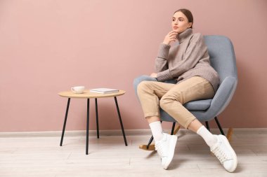 Güzel bir kadın, bej duvarın yanındaki sallanan sandalyede rahatlıyor.