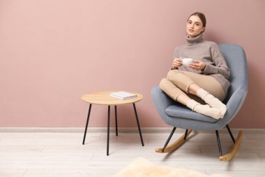 Beautiful woman with cup of drink relaxing in comfortable rocking chair near beige wall indoors, space for text