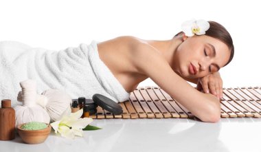 Güzel bir kadın beyaz arka planda bambu paspasın üzerinde yatıyor. Ayna yüzeyinde kaplıca ürünleri ve çiçekler