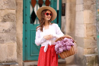 Şapkalı ve güneş gözlüklü gülümseyen kadın, bina yakınlarında bir sepet leylak çiçeğiyle.