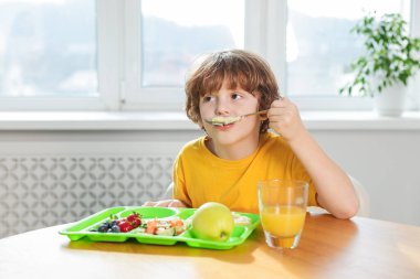 Okuldaki ahşap masada yemek yiyen şirin küçük çocuk.