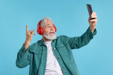 Kulaklıklı yaşlı bir adam açık mavi arka planda selfie çekiyor.