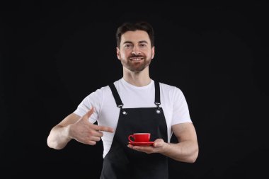 Siyah arka planda bir fincan sıcak içeceğe işaret eden gülümseyen baristanın portresi.