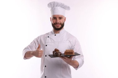 Üniformalı mutlu pastacı lezzetli çikolatalı tatlılar tutuyor ve beyaz arka planda başparmaklarını gösteriyor.