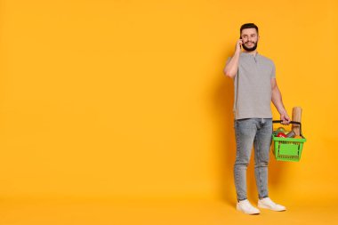 Alışveriş sepeti dolusu ürünü olan bir adam akıllı telefonuyla turuncu arka planda konuşuyor, mesaj için yer var.
