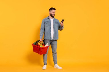 Ürünlerle dolu alışveriş sepeti ve turuncu arka planda akıllı telefonu olan adam.