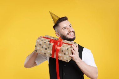 Doğum günün kutlu olsun. Turuncu arka planda parti şapkası ve hediye kutusu olan yakışıklı bir adam.