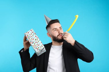 Doğum günün kutlu olsun. Parti şapkası, üflemeli ve açık mavi arka planda hediye kutusu olan yakışıklı bir adam.