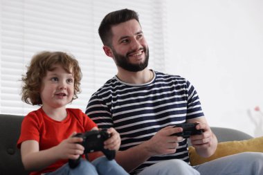 Mutlu baba ve oğlu evdeki kanepede oyun kumandalarıyla oynuyorlar.