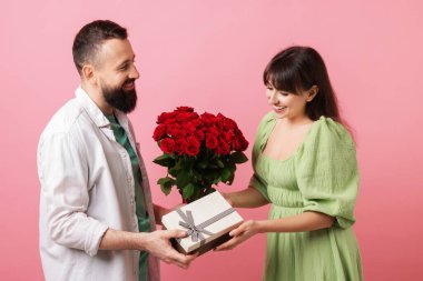Kırmızı gül buketini ve hediye kutusunu pembe arka plandaki sevgilisine sunan bir adam.
