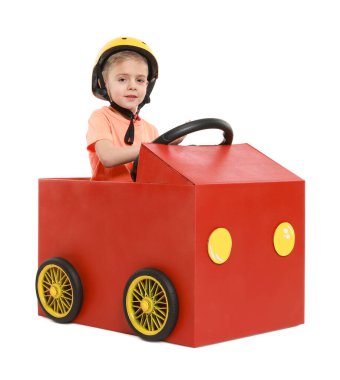 Küçük çocuk beyaz arka planda kartondan yapılmış araba kullanıyor.