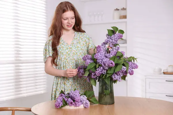 Gülümseyen genç kız, iç mekandaki masada vazoya leylak çiçekleri koyuyor.