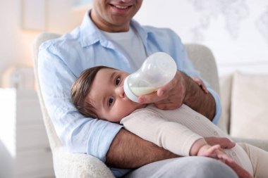 Babası küçük bebeğini evdeki koltukta biberonla besliyor.