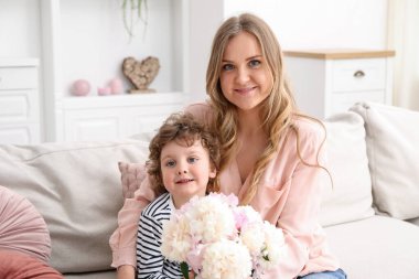 Küçük çocuk evdeki kanepede annesine güzel şakayık çiçekleri veriyor.