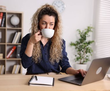 Bir fincan sıcak içecekle ev ofisinin masasında çalışan bir kadın.