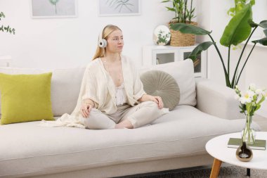 Feng shui. Kulaklıklı genç bir kadın evdeki kanepede meditasyon yapıyor.