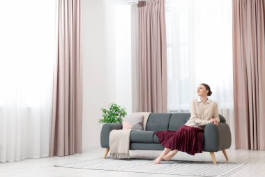 Kadın pencerenin yanındaki kanepede oturuyor ve evinde şık perdeler var. Metin için boşluk