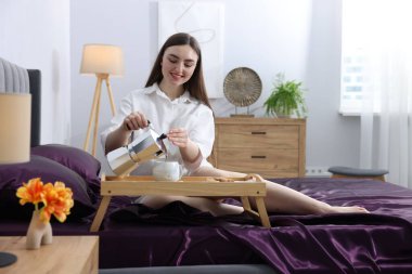 Gülümseyen kadın, kahvesini Moka pot 'tan bardağa dolduruyor. Yatakta mor ipek çarşaflar var.