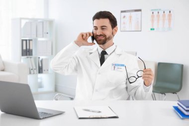 Gözlüklü profesyonel doktor hastanedeki beyaz masada akıllı telefondan konuşuyor.