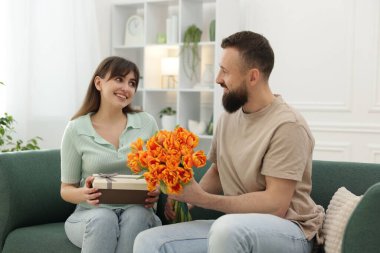 Bir adam evdeki kanepede sevgili karısına güzel laleler sunuyor.