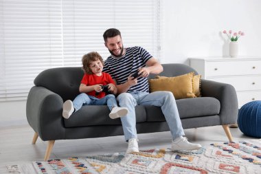 Mutlu baba ve oğlu evdeki kanepede oyun kumandalarıyla oynuyorlar.