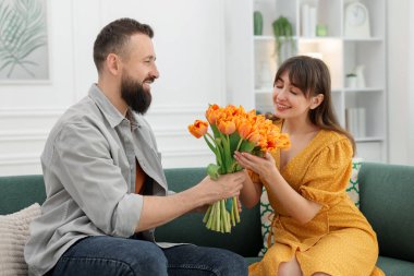 Bir adam evdeki kanepede sevgili karısına güzel laleler sunuyor.