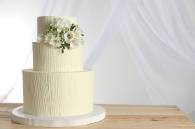 Ahşap masadaki çiçek desenli nefis düğün pastası ve beyaz arka plandaki perdeler.