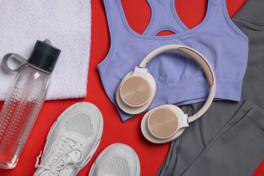 Spor kıyafetleri, spor ayakkabıları, havlular, kulaklıklar ve su şişesi kırmızı arka planda.