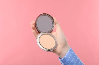 Kadının elinde açık yüz pudrası, pembe arka planda ayna, yakın plan.