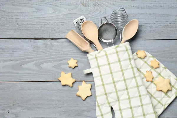 Fırın eldiveninde farklı mutfak aletleri ve açık gri ahşap masada yıldız şeklinde kurabiyeler. Metin için boşluk