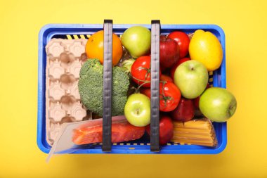 Sarı arka planda ürünlerle dolu plastik alışveriş sepeti.