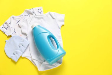 Bir şişe yumuşatıcı ve sarı arka planda bebek kıyafetleri. Metin için boşluk