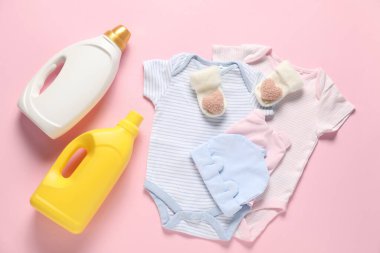 Yumuşatıcı şişeler ve pembe arka planda bebek kıyafetleri.