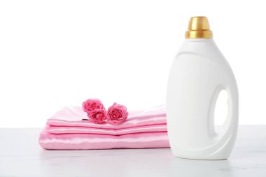 Bir şişe yumuşatıcı, beyaz arkaplan karşısında üst üste yığılmış giysiler ve çiçekler.