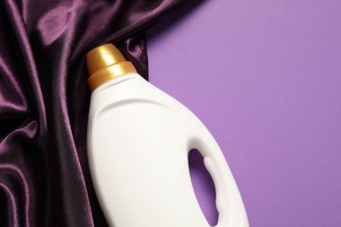 Bir şişe yumuşatıcı ve mor arka planda renkli kumaş, üst manzara. Metin için boşluk
