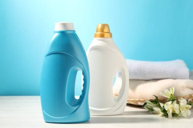 Yumuşatıcı şişeler, temiz kıyafetler ve açık mavi arka planda beyaz ahşap masa üzerinde zambak çiçekleri.