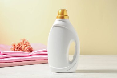 Bir şişe yumuşatıcı, temiz kıyafetler ve karanfil çiçekleri açık sarı arka planda beyaz ahşap masa üzerinde