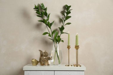 Zarif şamdanlar, yanan mumlar, vazoda dallar ve bej duvarın yanındaki şifoniyerde diğer dekorlar.