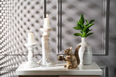 Zarif şamdanlar, yanan mumlar, vazoda dallar ve gri duvarın yanındaki şifoniyerde diğer dekorlar.