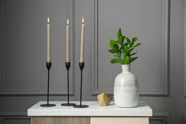 Zarif şamdanlar, yanan mumlar, vazoda dallar ve gri duvarın yanındaki şifoniyerde diğer dekorlar.