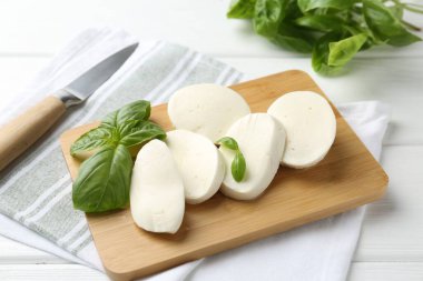 Lezzetli mozzarella peyniri parçaları, fesleğen yaprakları ve beyaz ahşap masa üzerinde bıçak, yakın plan.