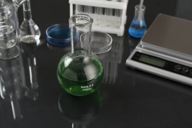 Yeşil sıvılı matara, diğer laboratuvar camları ve ölçekli siyah ayna masası, yakın plan.