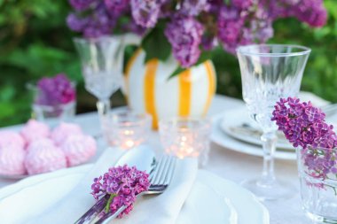 Güzel leylak çiçekleri, zephyrler, tabaklar, çatal bıçaklar, bardaklar ve yanan mumlar bahçede, yakın plan