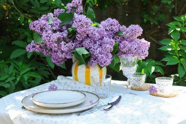 Bahçedeki masada güzel leylak çiçekleri, tabaklar, çatal bıçaklar, bardaklar ve mumlar