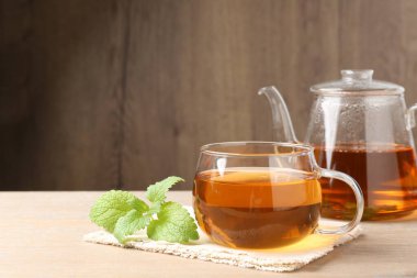 Cam fincanda aromatik limon merhemi çayı, çaydanlık ve ahşap masada taze yapraklar, metin için yer.